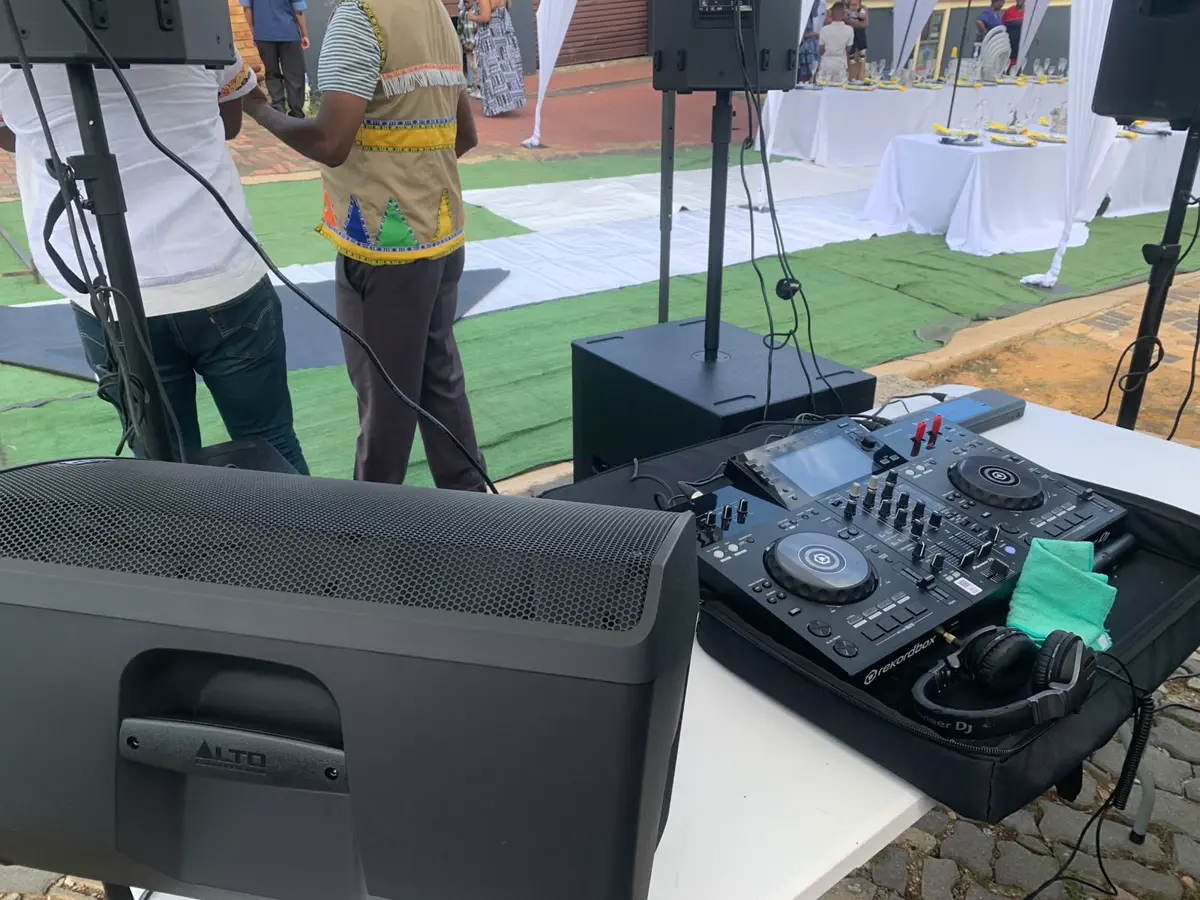 Outdoor DJ setup with Alto speaker and Pioneer DJ controller, people and decorated tables in the background
